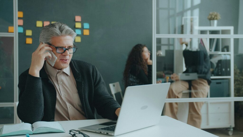 B2B-Inkasso im Alltag: Geschäftsmann telefoniert am Laptop in einem Büro mit Kollegen im Hintergrund