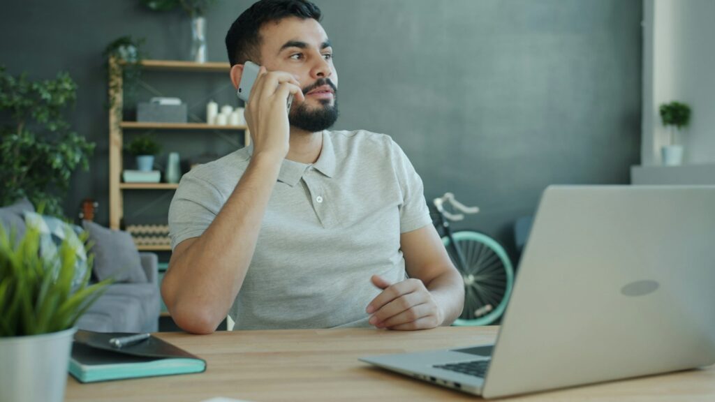 Deepfake-Betrug per Telefon: Mann telefoniert am Schreibtisch mit dem Smartphone vor seinem Laptop